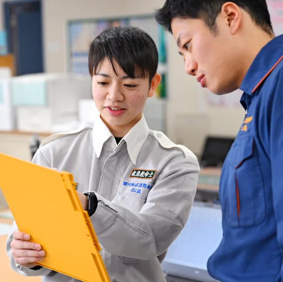 松浪さんが手に持った黄色いクリップボードを指差し、クリップボードを覗き込んでいる消防隊の人に説明している様子