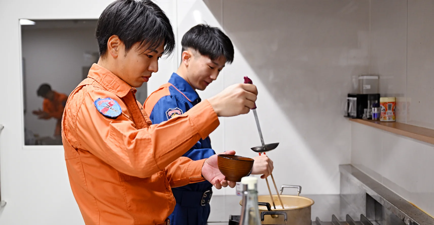 救助隊の方がだしをお椀によそい、奥にいる消防隊の人が鍋で麺を茹でている様子